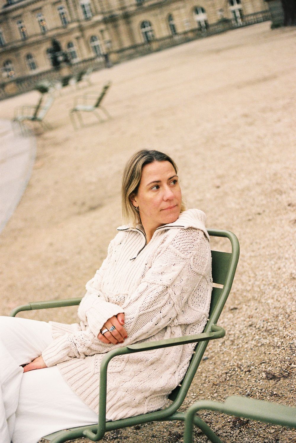 Someone in a cream cable knit sweater sits in a green metal chair at a grand European palace garden.