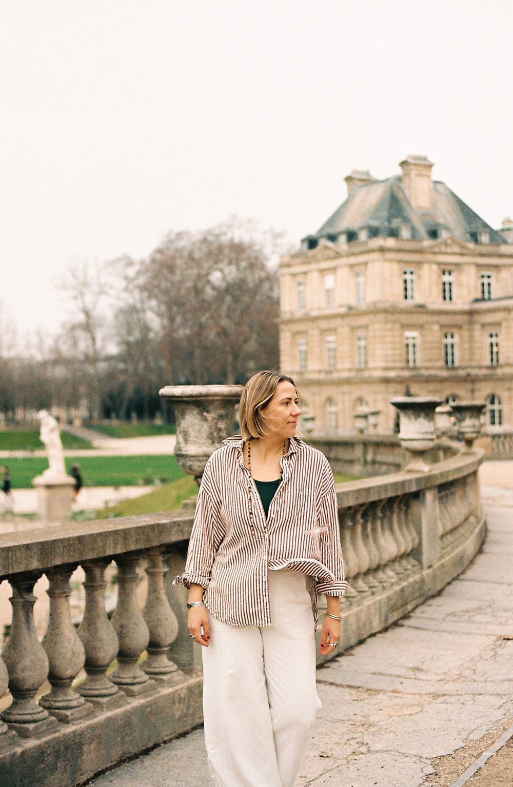 A person in a striped shirt and white pants stands on a balcony overlooking the Luxembourg Gardens in Paris.