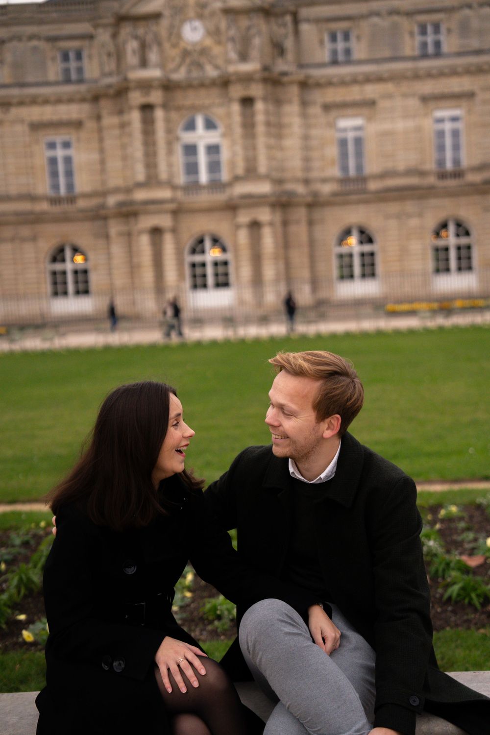 A couple shares a joyful moment sitting together on a bench outside an elegant historic building with arched windows.