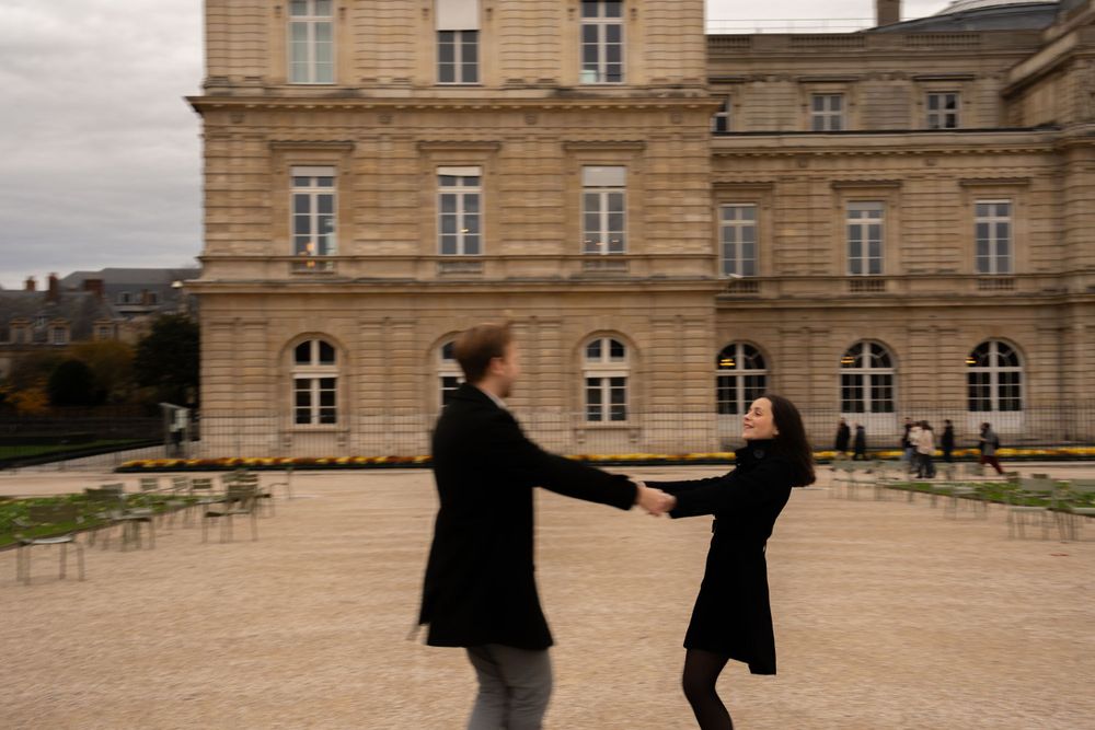 Two people in dark coats hold hands and dance together in front of a grand historic French palace.