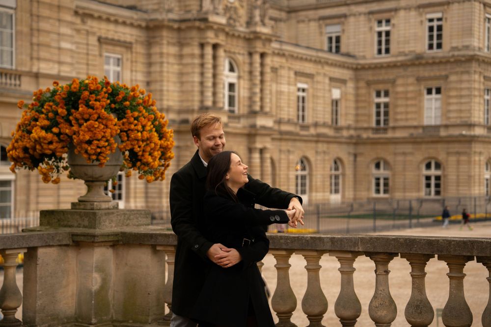 A couple in dark coats stands on an elegant balcony with orange flowers near a historic limestone palace building.