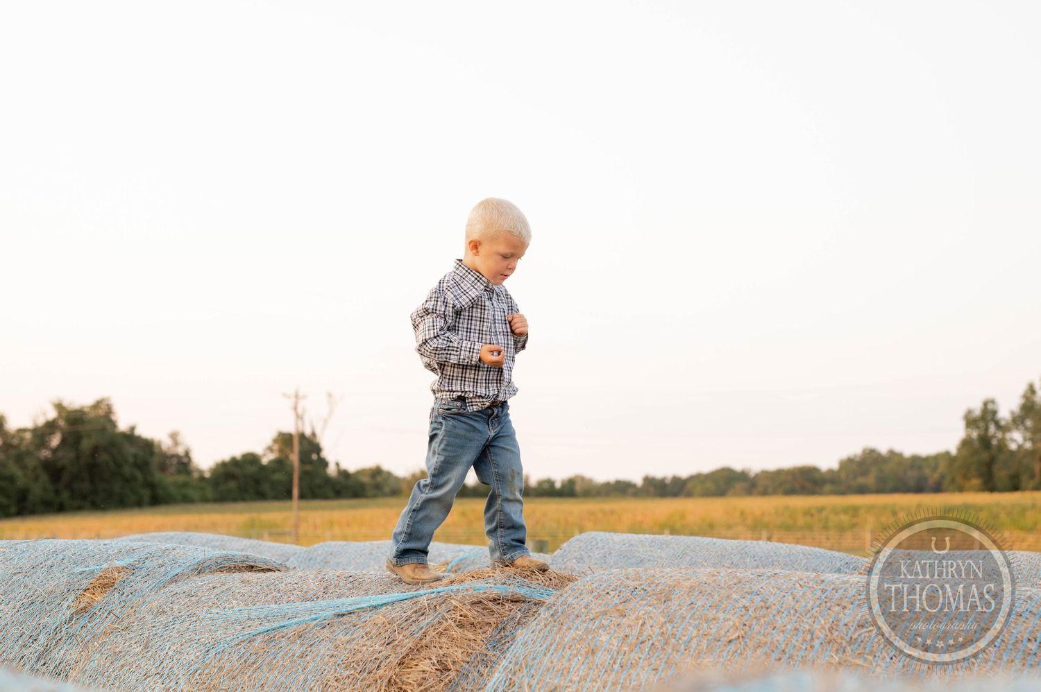 Small child in western wear stands on large round hay bale in field.