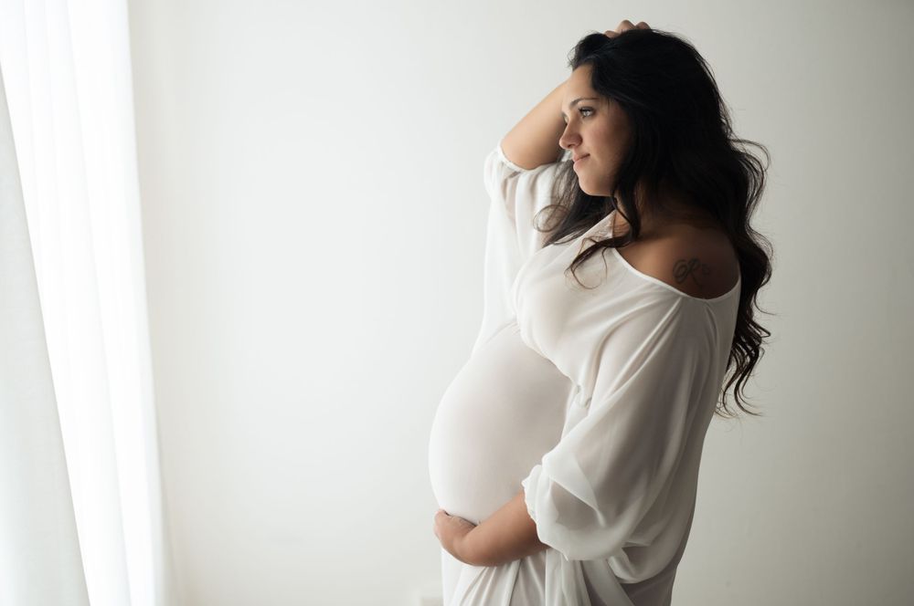 Giovane donna incinta con abito bianco, con una mano sotto il pancione e con l'altra si accarezza i capelli