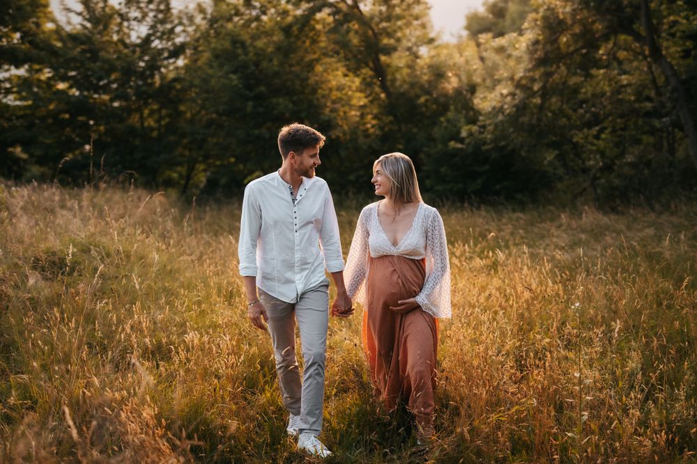 Coppia di futuri genitori vestiti per un servizio fotografico che cammina in mezzo a un prato circondati dalla natura verde di luglio