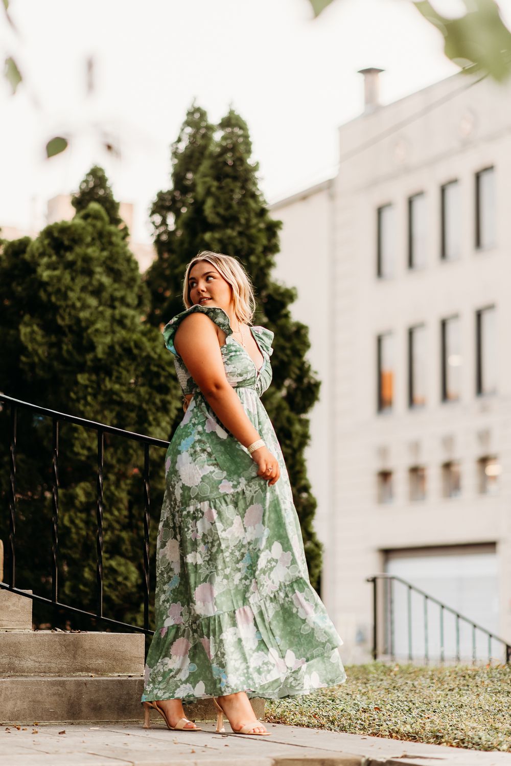 Model in flowing green and white floral maxi dress poses on stone steps near a white building.