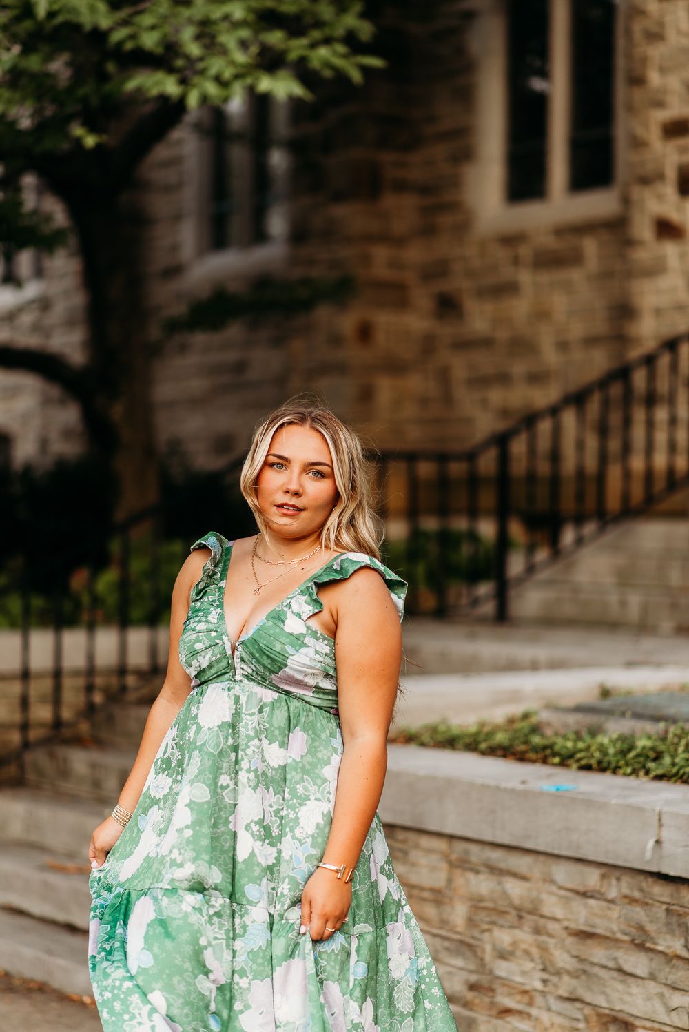 Woman in green floral maxi dress poses outdoors near stone building steps.