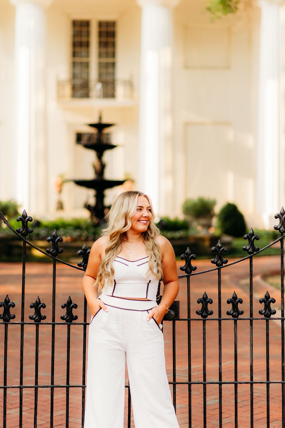 Fashion portrait of a white jumpsuit outfit with black iron gates and fountain in architectural background.