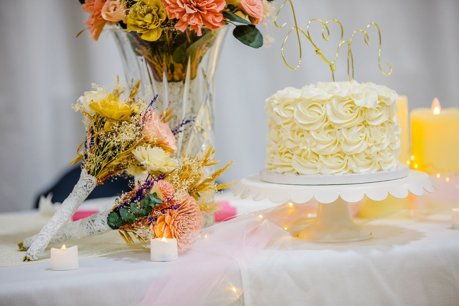 White frosted wedding cake with swirl pattern sits beside colorful floral arrangement and glowing yellow candle.