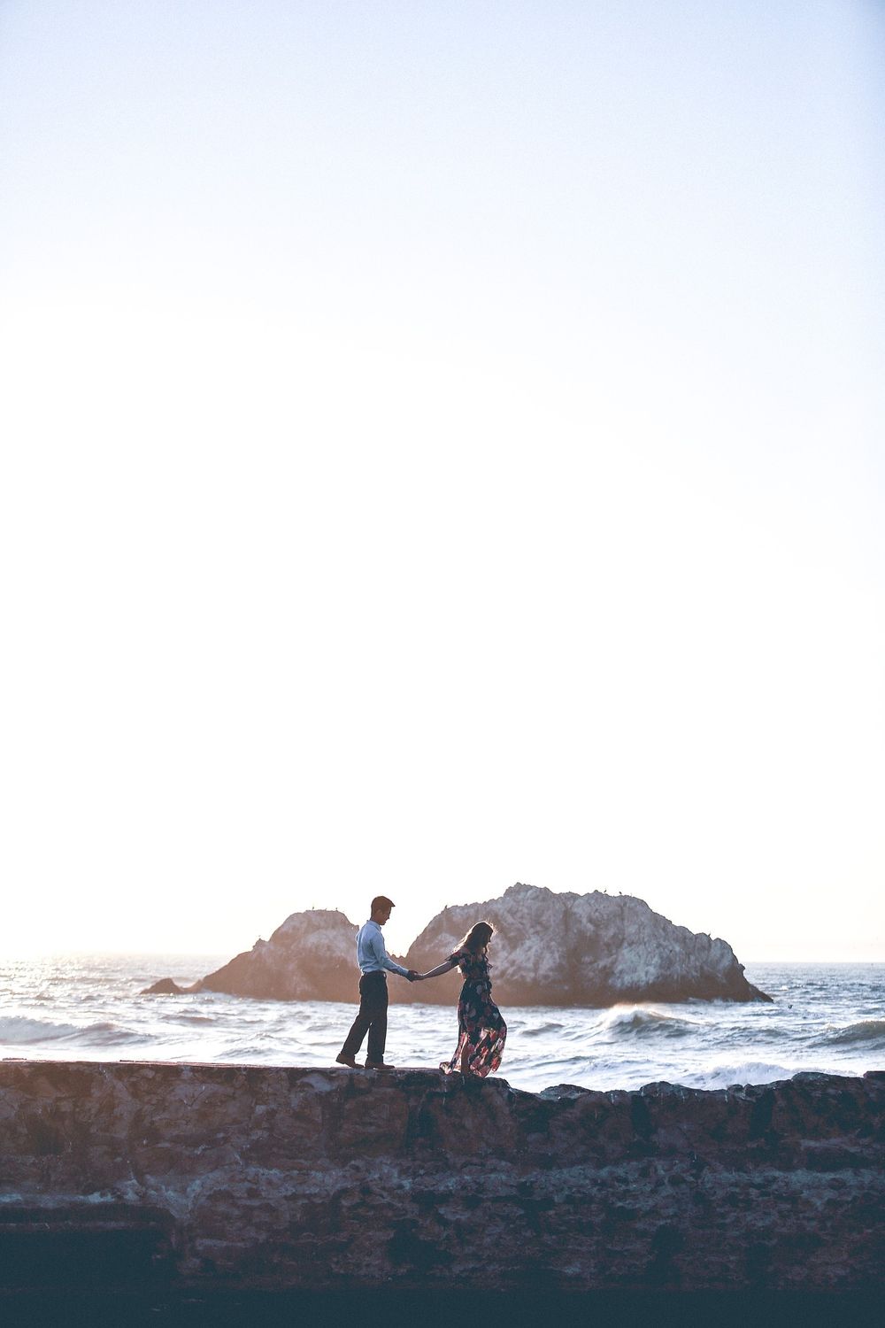 Silhouettes of couple standing on rocky cliff overlooking ocean at sunset.