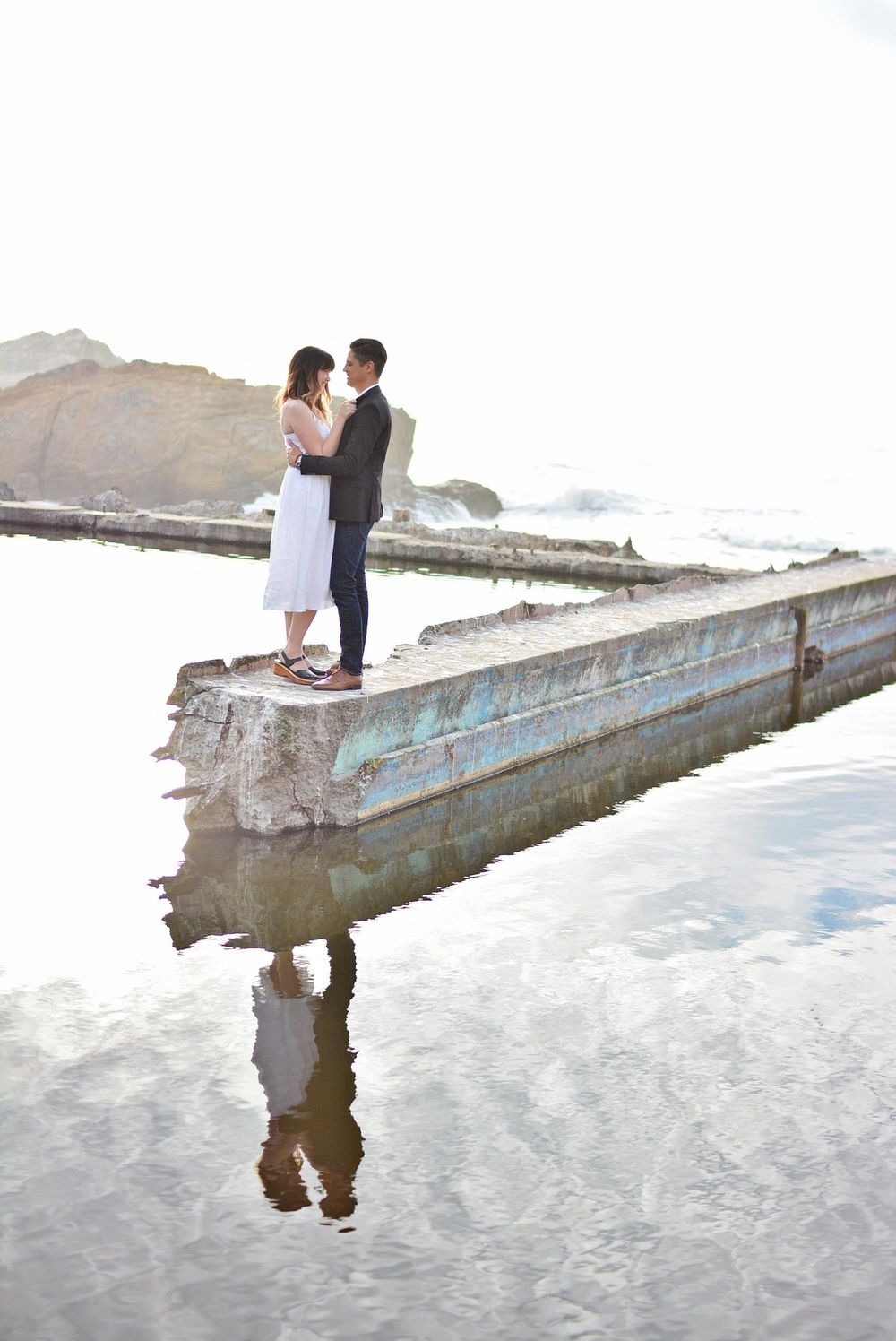 Couple standing on concrete pier with water reflections during seaside photoshoot.