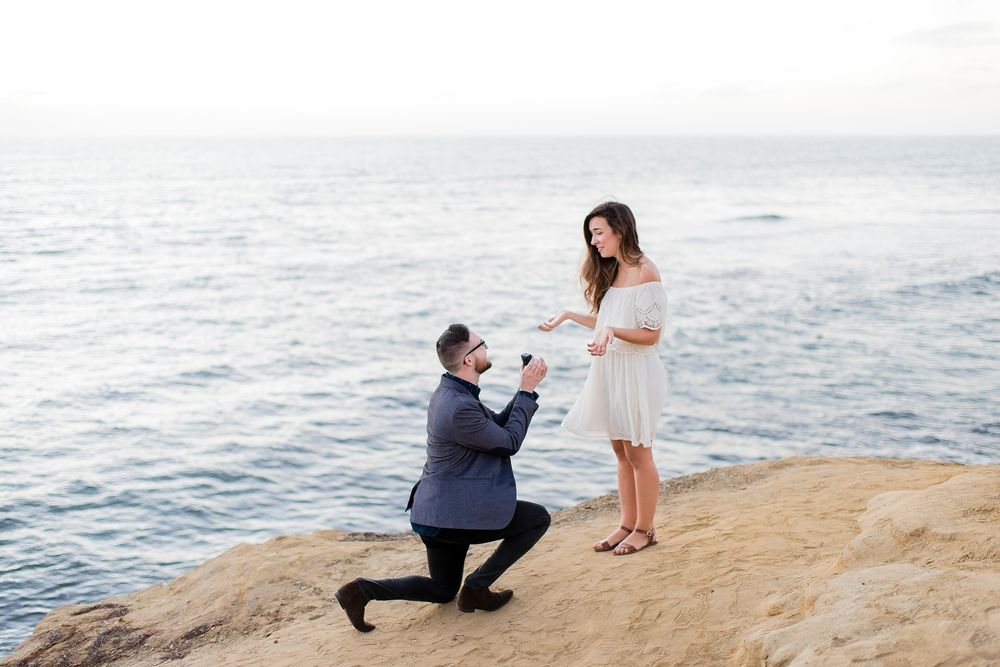 Couple sharing a romantic moment on a rocky beach at sunset with ocean waves in the background.