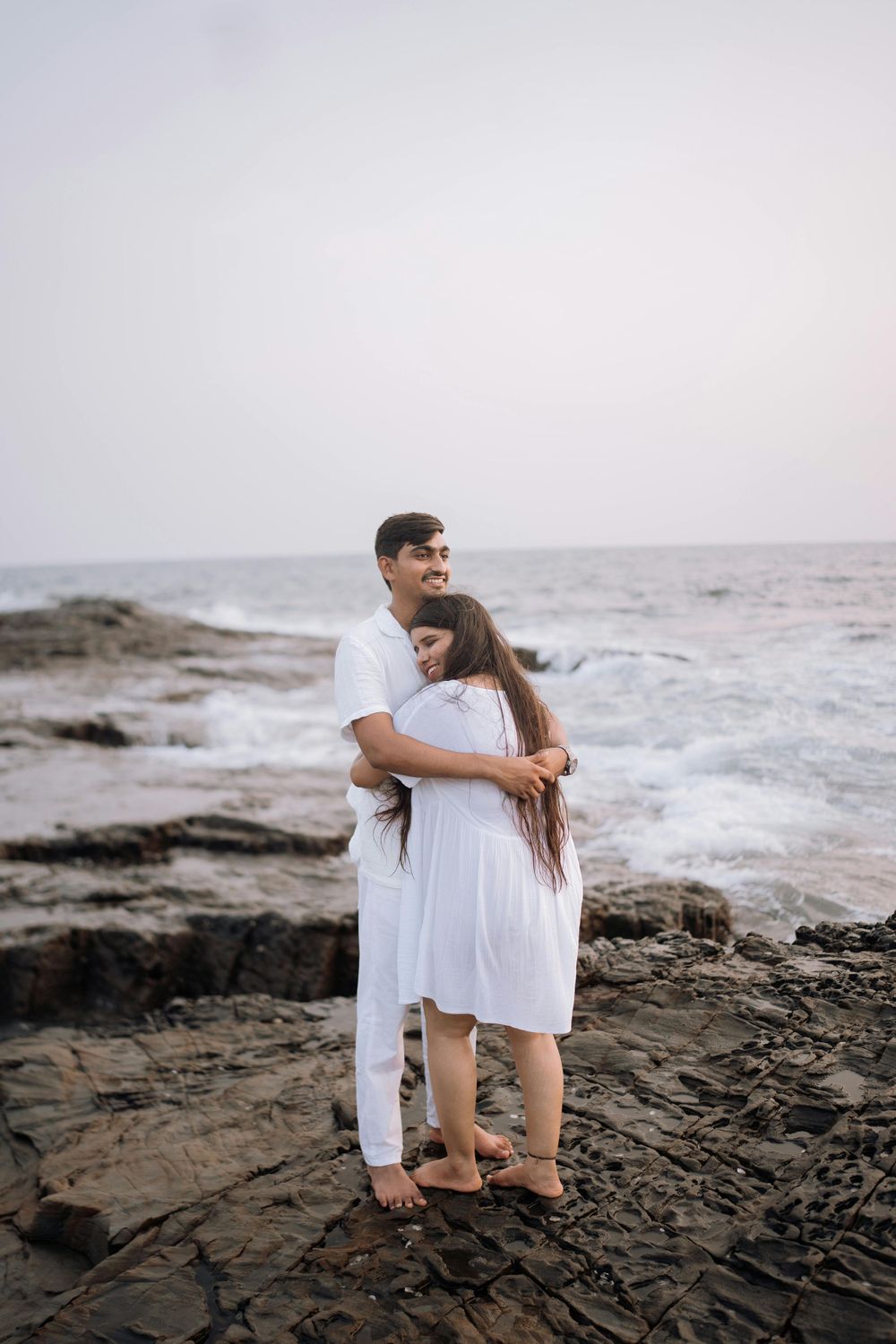 Couple in white outfits embracing on rocky beach shoreline during cloudy day.