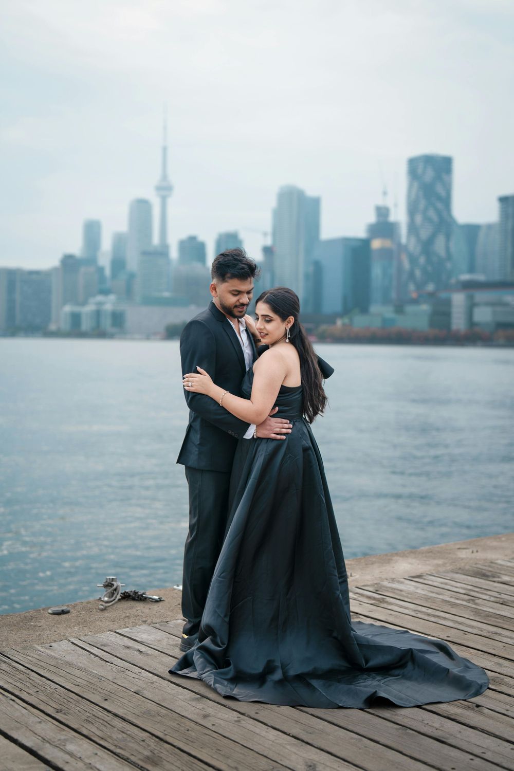 Couple in formal attire posing on wooden pier with Toronto skyline visible across the water.