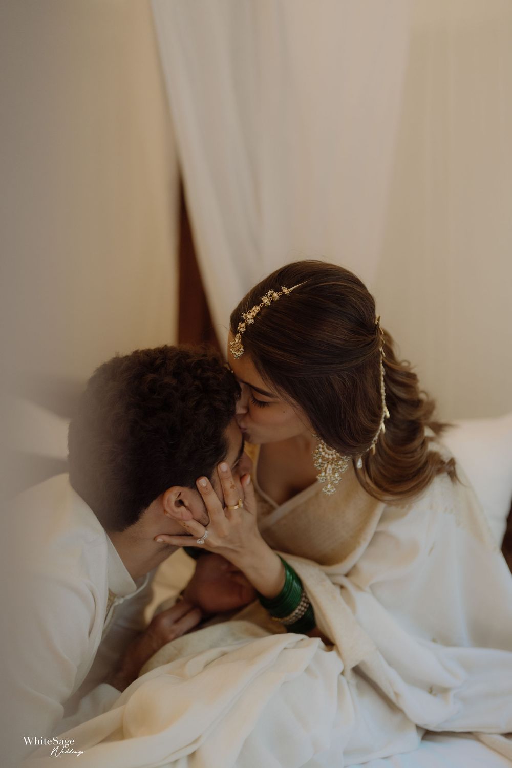 A romantic wedding moment captured in soft lighting as a couple shares an intimate kiss draped in white fabric.