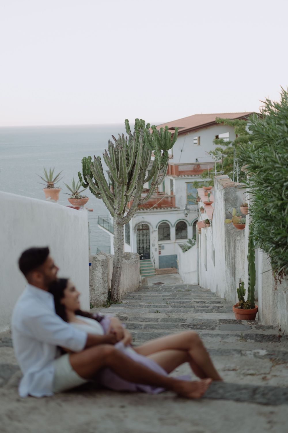 A couple relaxes on steps along a charming Mediterranean street with white walls, arched doorways and potted cacti.