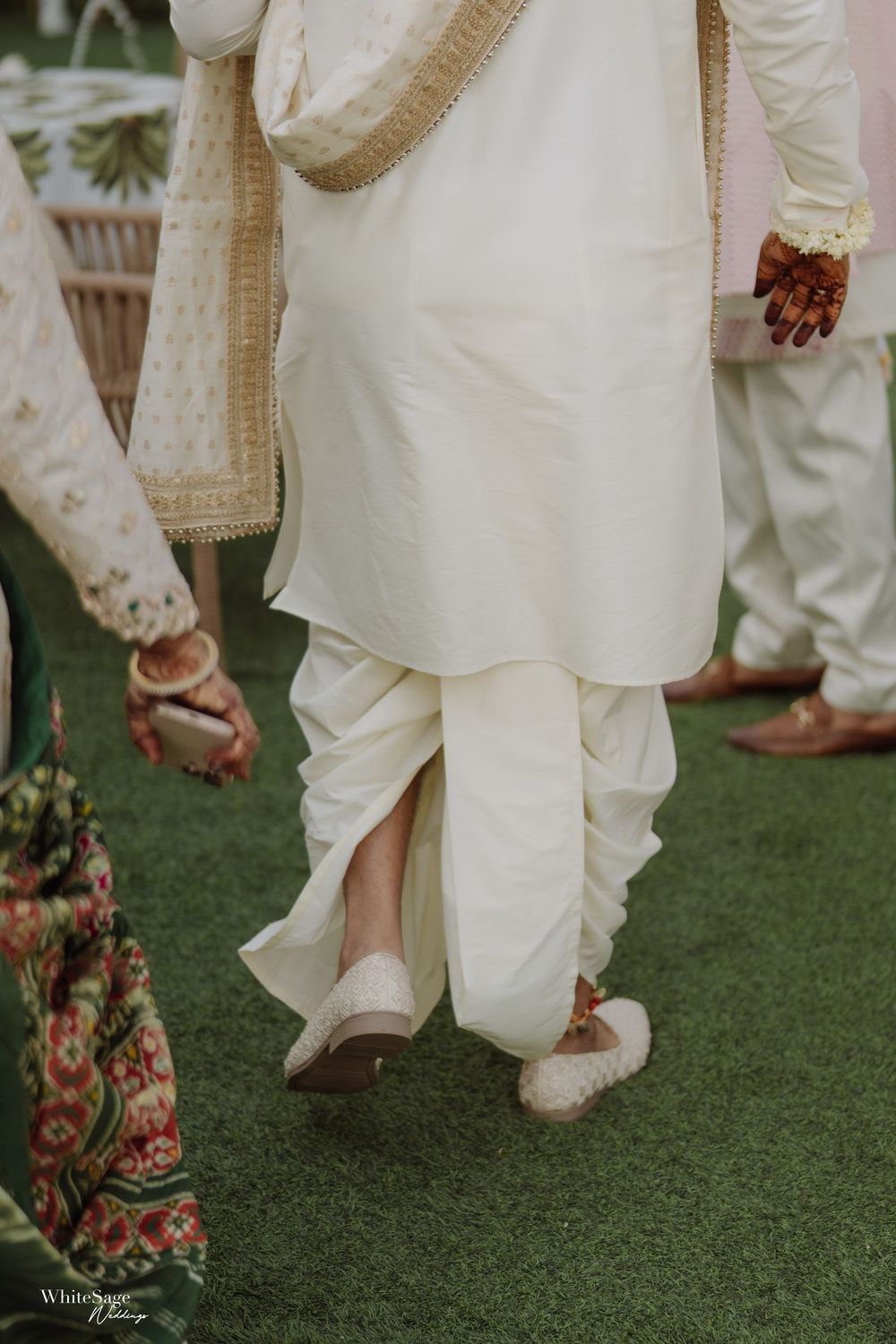 Close-up view of white traditional Indian wedding attire and ornate footwear on grass during a ceremony.