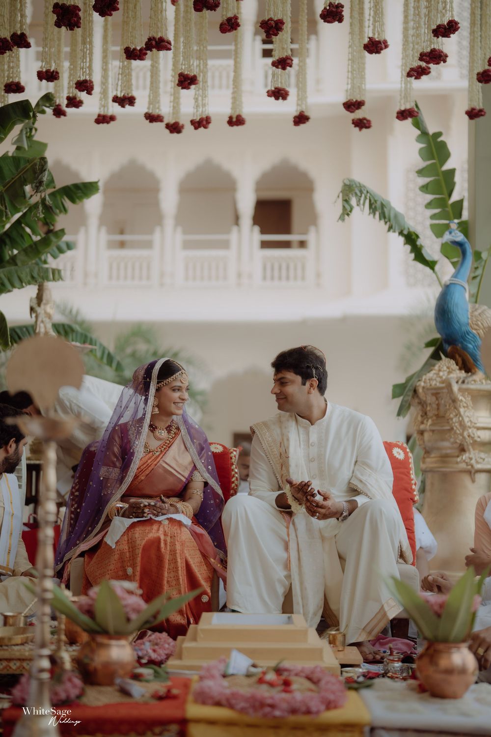Traditional Indian wedding ceremony with red floral decorations hanging from above and tropical greenery in an ornate setting.