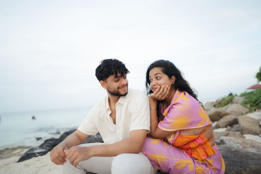 A romantic couple embraces on a scenic rocky beach during an overcast day with ocean waves in the background.