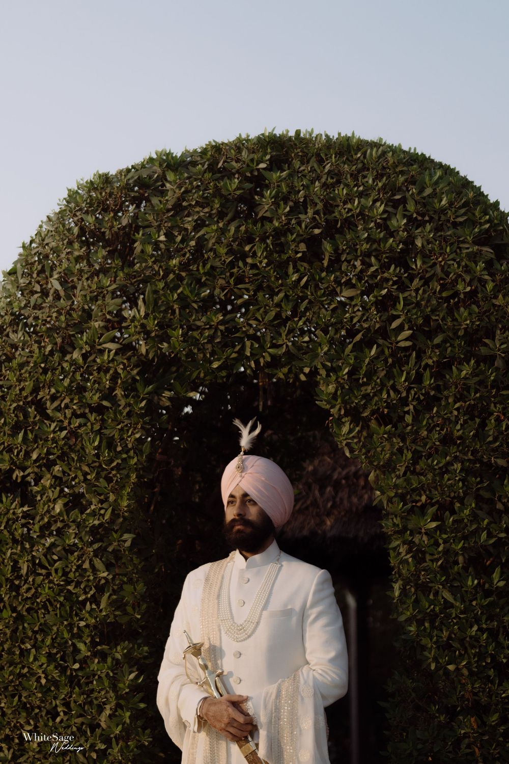 Groom in traditional white attire and turban standing against a meticulously manicured round hedge backdrop.
