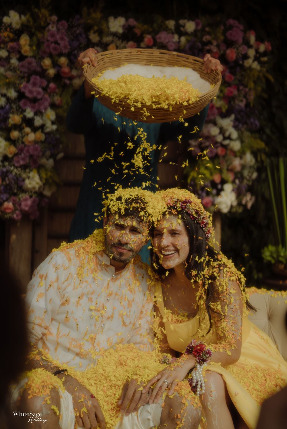Couple sits together under shower of yellow flower petals during festive haldi ceremony with floral decorations.