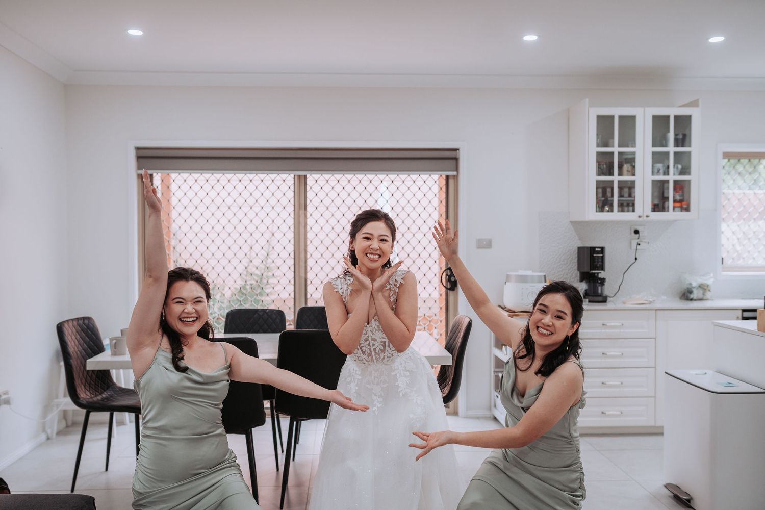 Three people celebrate joyfully in a modern white kitchen while wearing formal attire.
