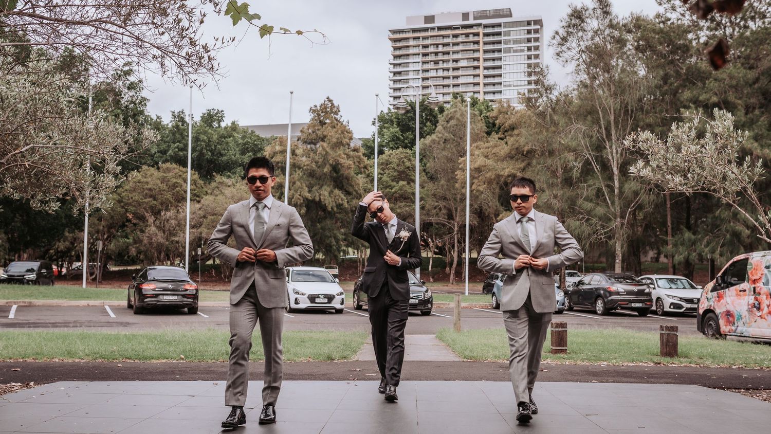 Three figures walk down a tree-lined urban street with a high-rise building visible in the distance.