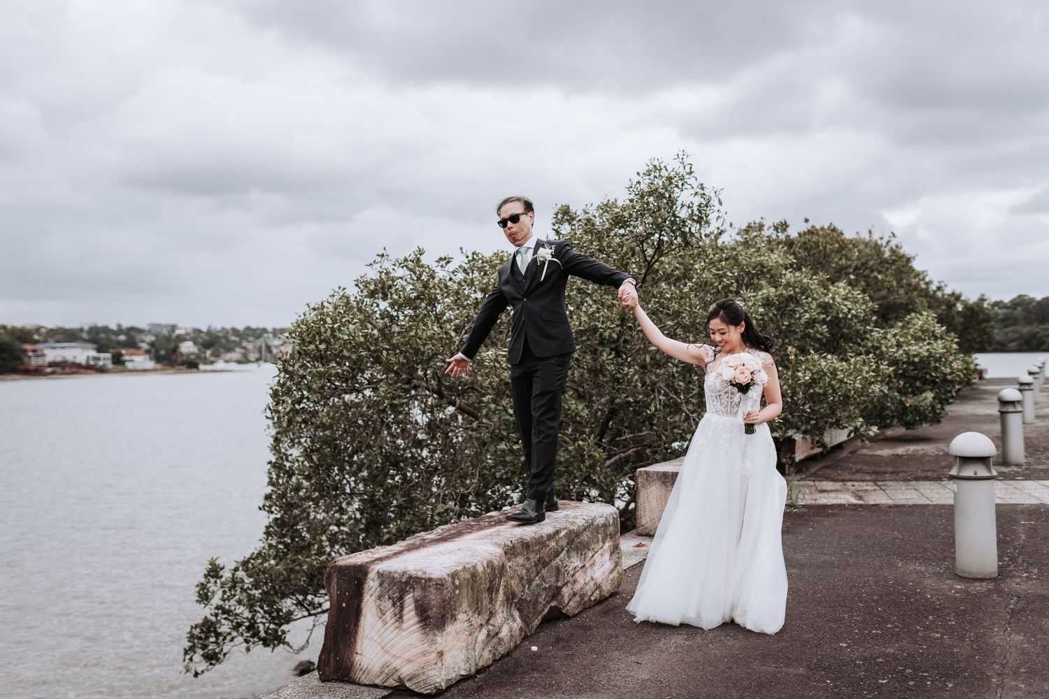 A couple in wedding attire poses playfully on a concrete wall by a waterfront.