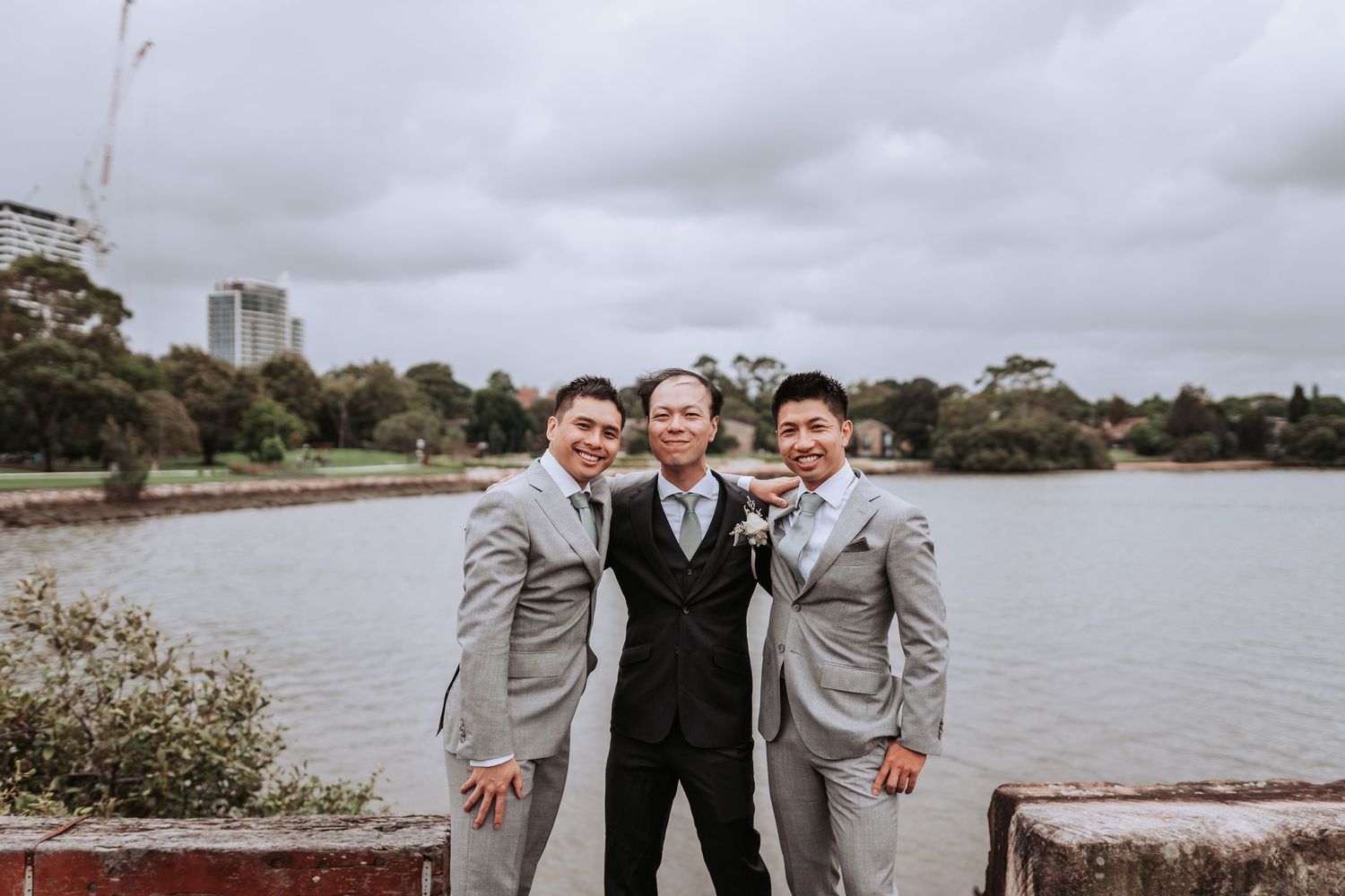 Three groomsmen in grey and black suits pose by a lake on an overcast day.