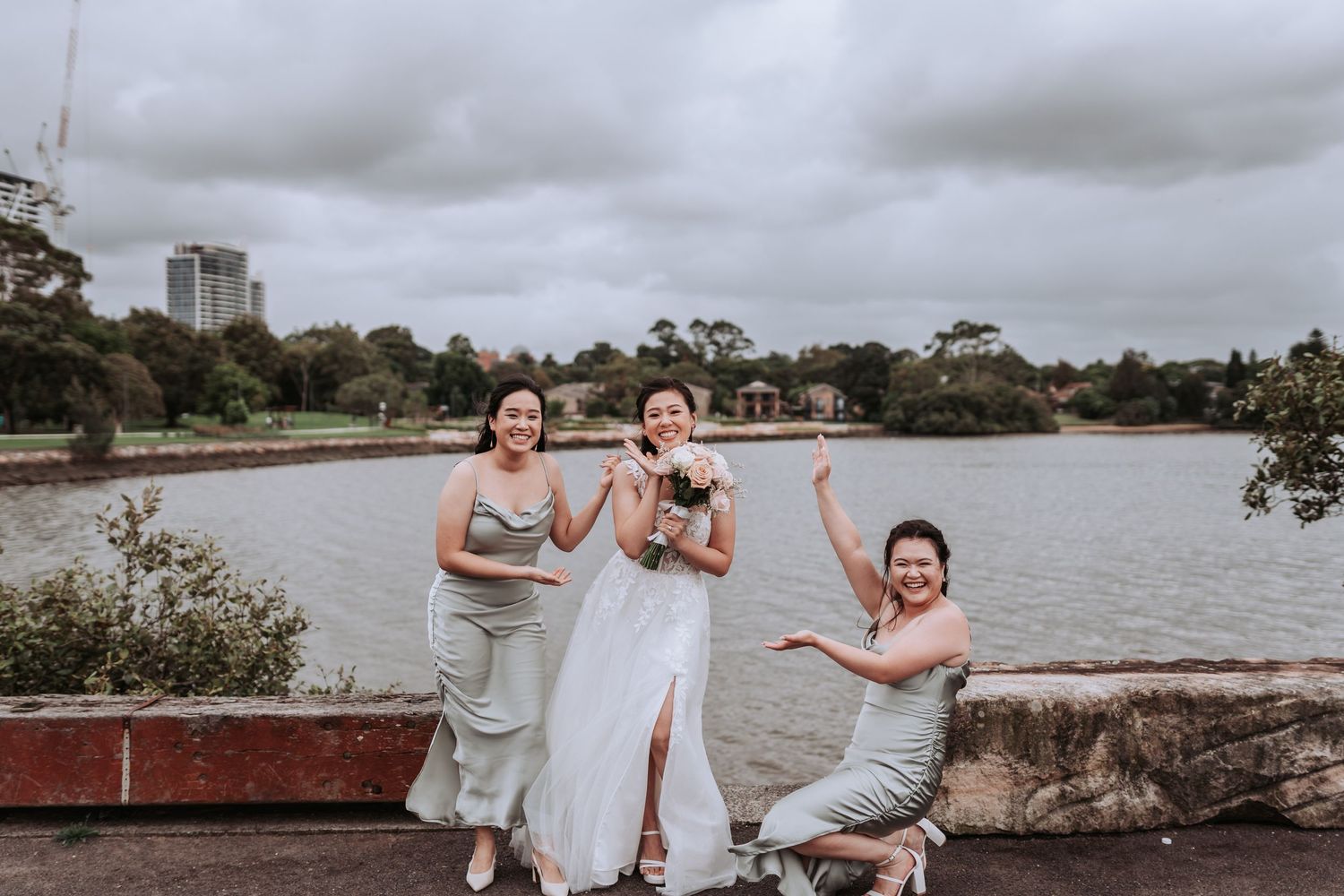 Three people in formal attire pose playfully by a waterfront on a cloudy day.