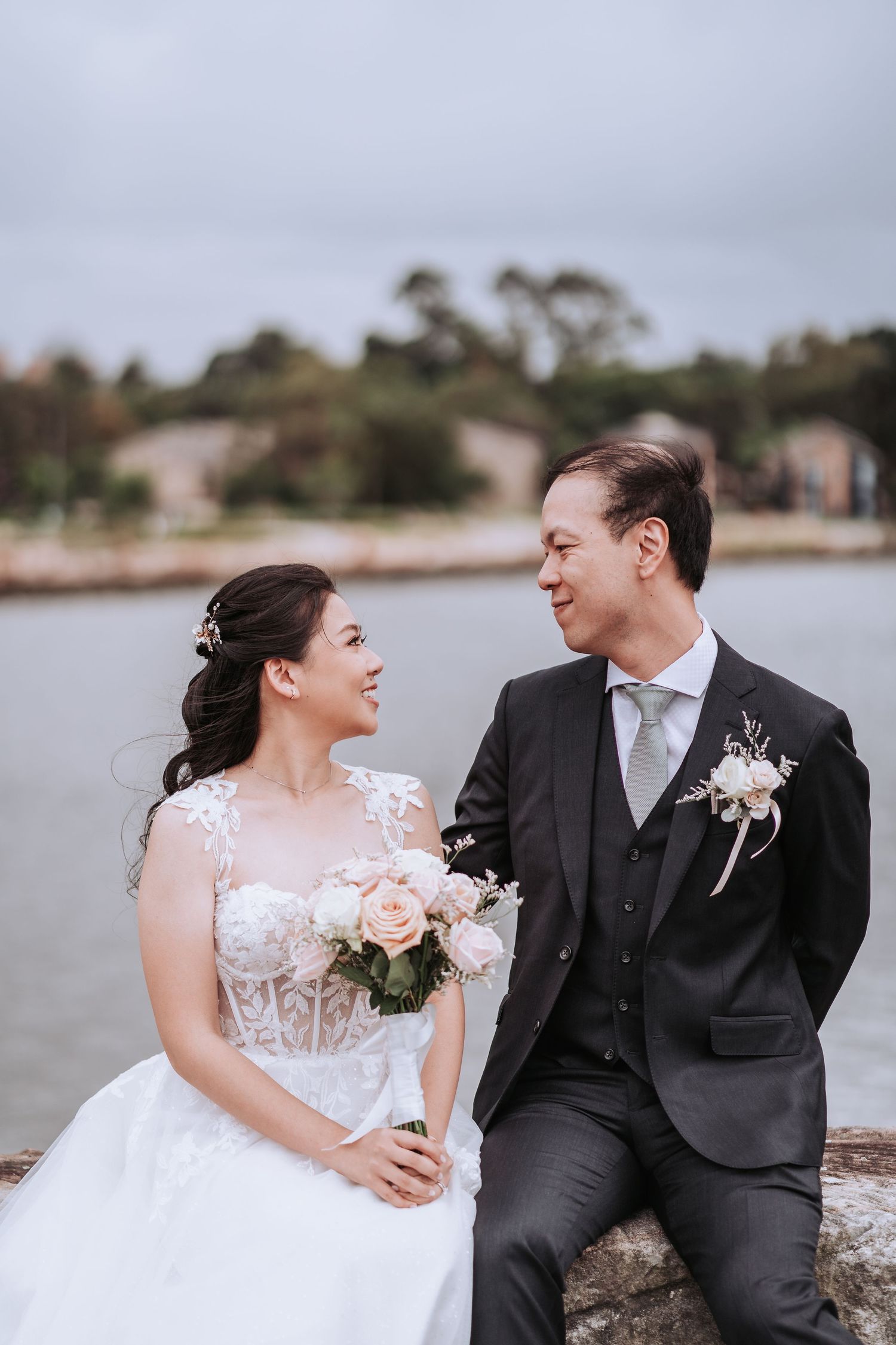 A romantic wedding couple sitting by a lake with overcast skies in the background.