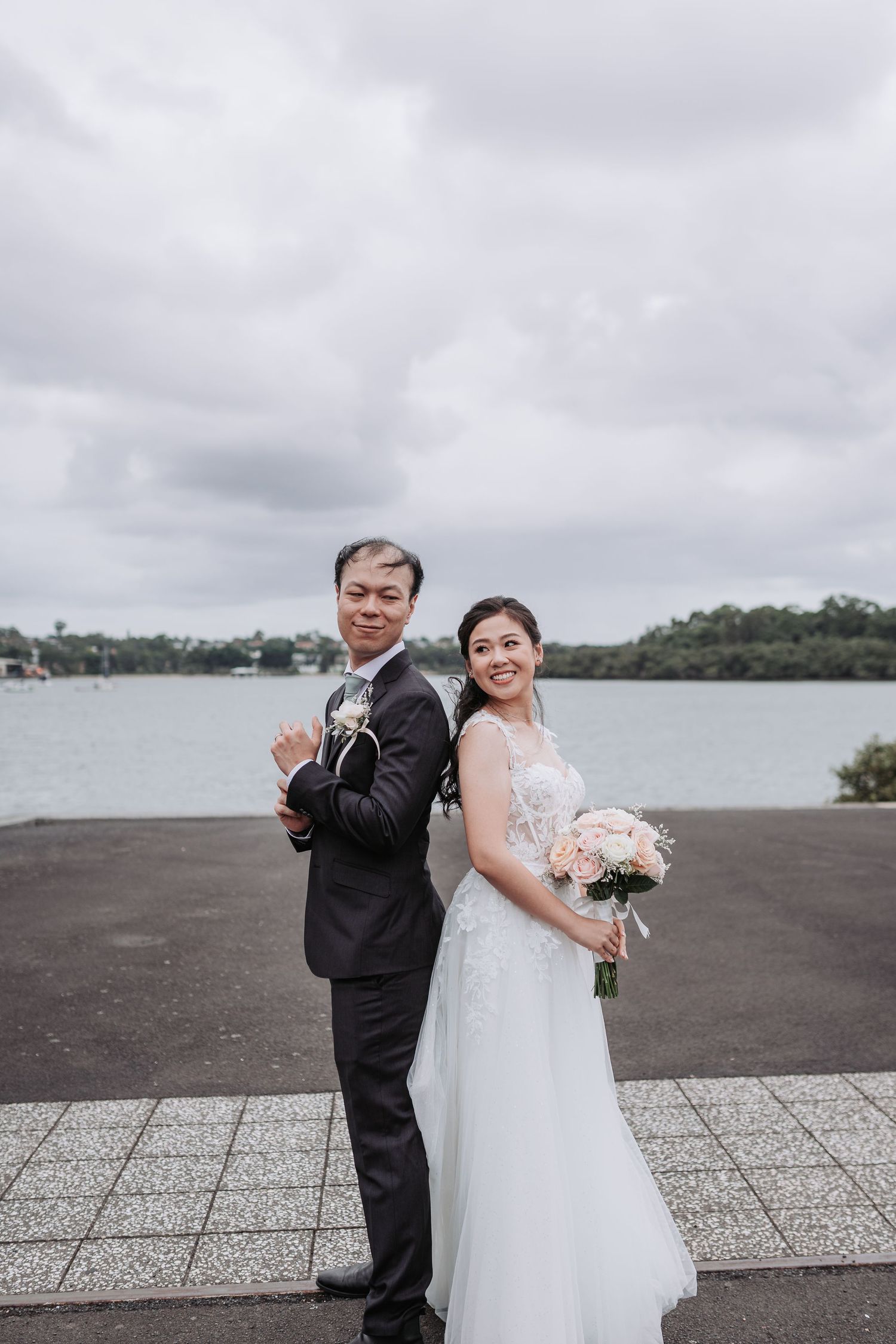 A couple in wedding attire pose back-to-back at a waterfront location under cloudy skies.