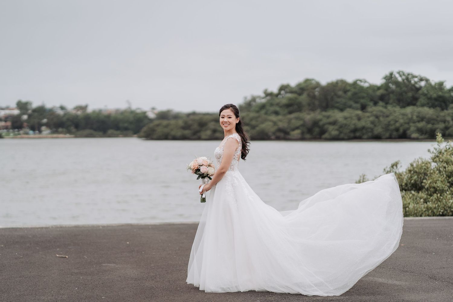 A bride in a flowing white dress poses by a lake with her dress billowing in the wind.
