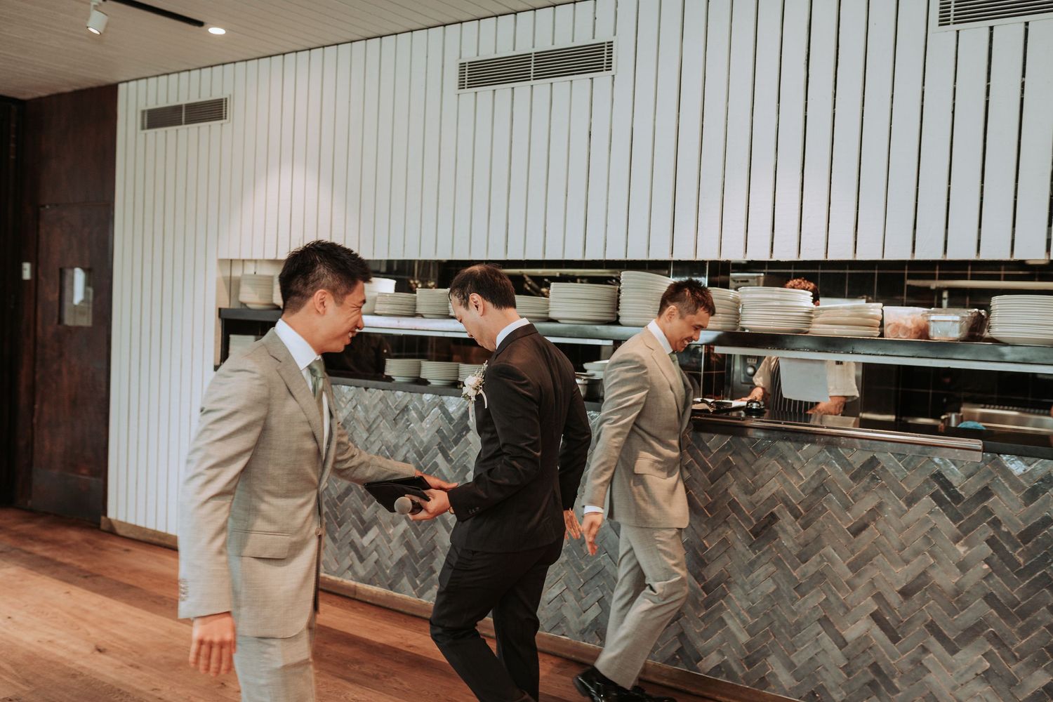 Three people in grey suits walk through a modern restaurant interior with herringbone tile walls.