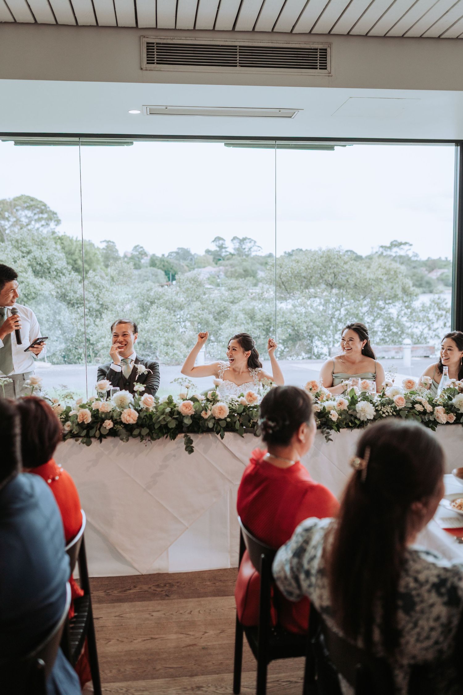 Wedding reception in a modern venue with floor-to-ceiling windows overlooking trees and greenery.