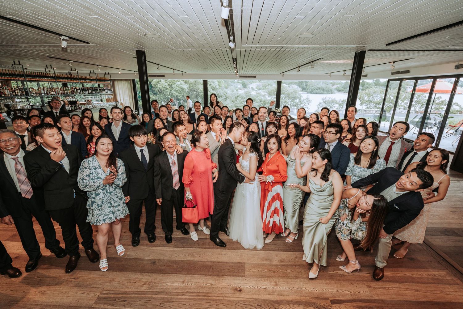 Large group photo of wedding guests gathered in an indoor reception venue with wooden floors.