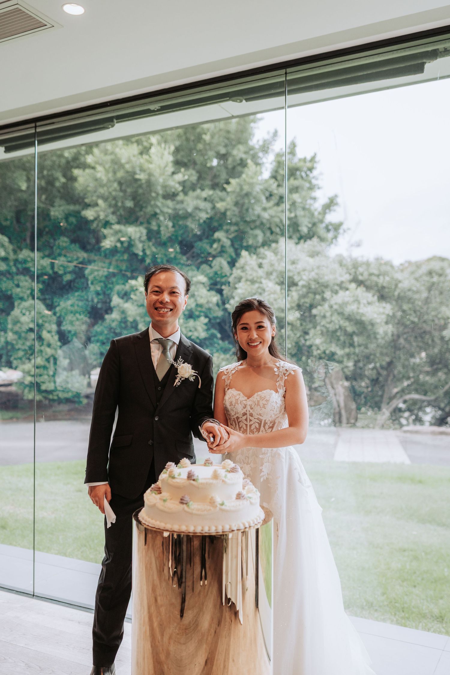 A wedding couple stands beside their elegant white wedding cake in a glass-walled venue.