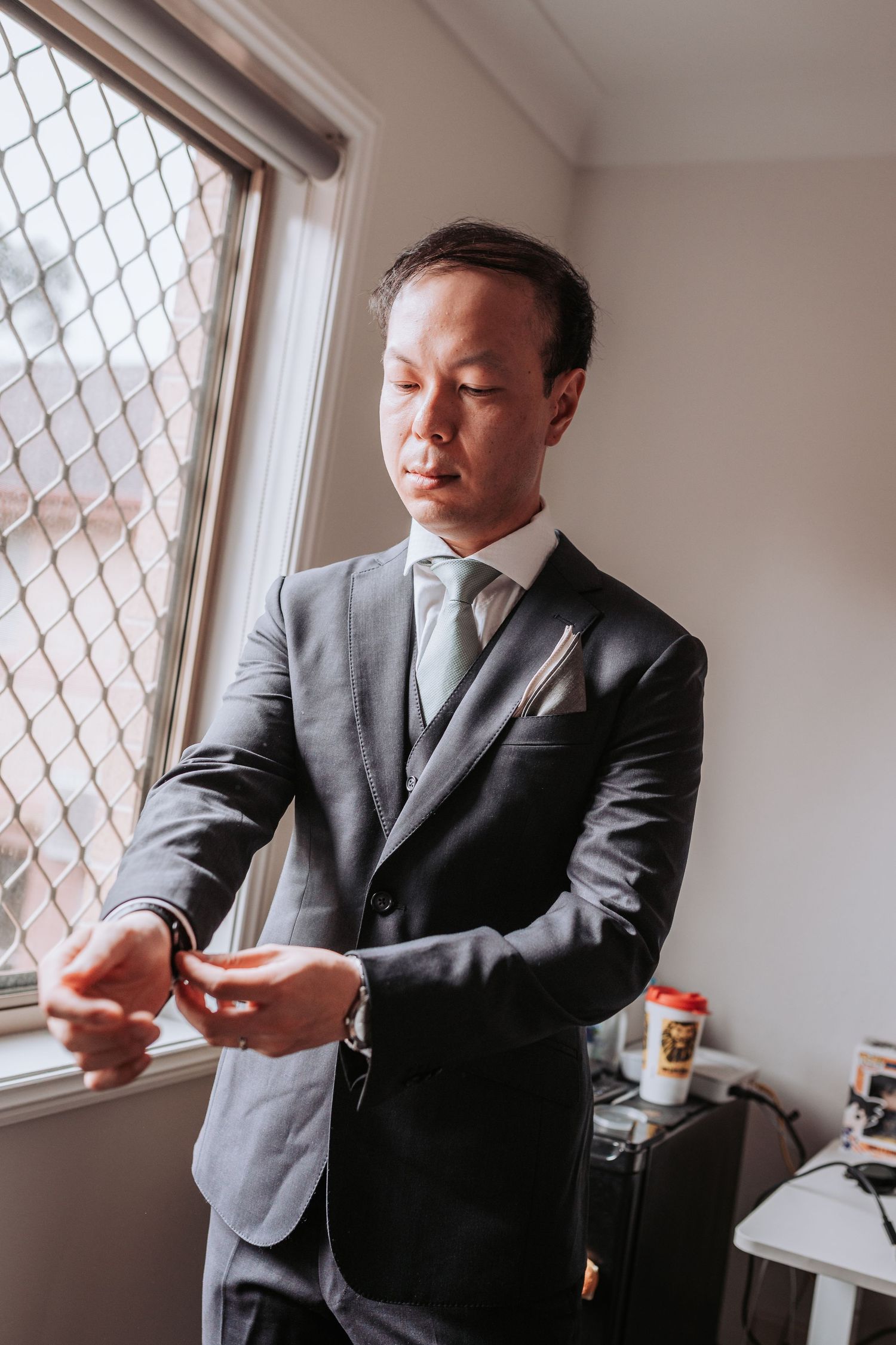 A person in a formal grey suit adjusts their tie while standing near a window with mesh security screen.