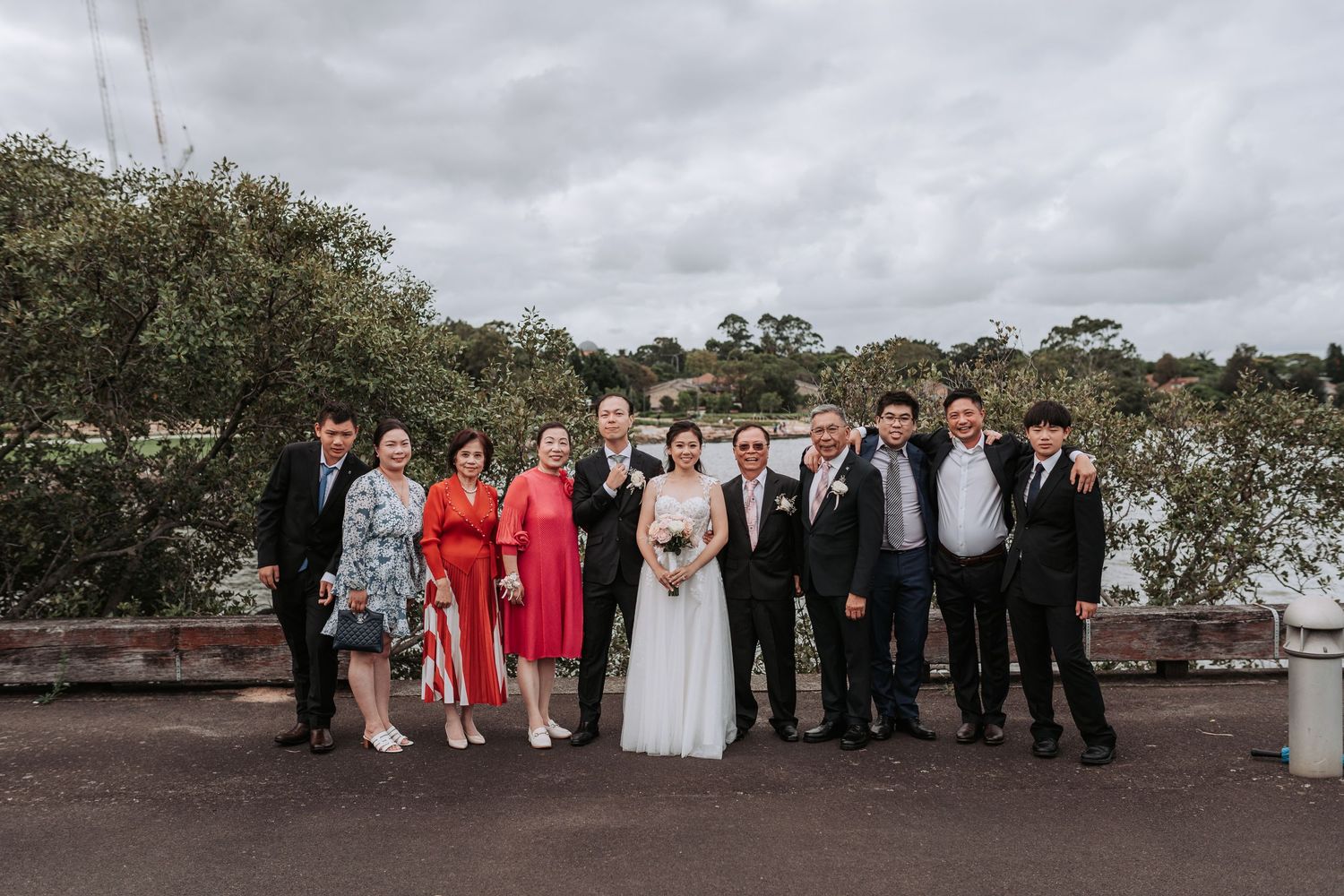 A wedding party poses for a group photo outdoors under cloudy skies with trees in the background.