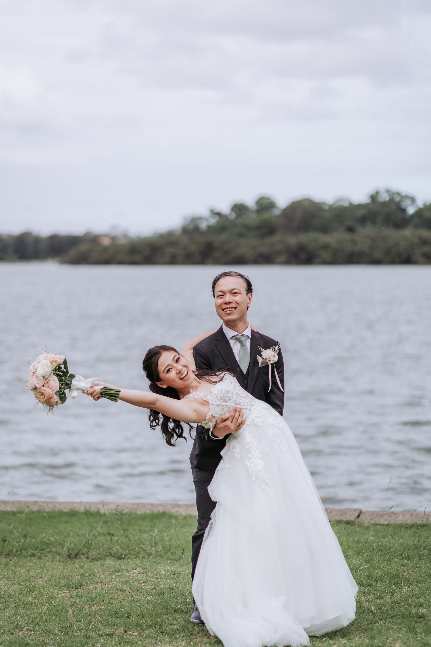 Romantic dip pose by newlyweds on lakeside lawn with cloudy sky backdrop.
