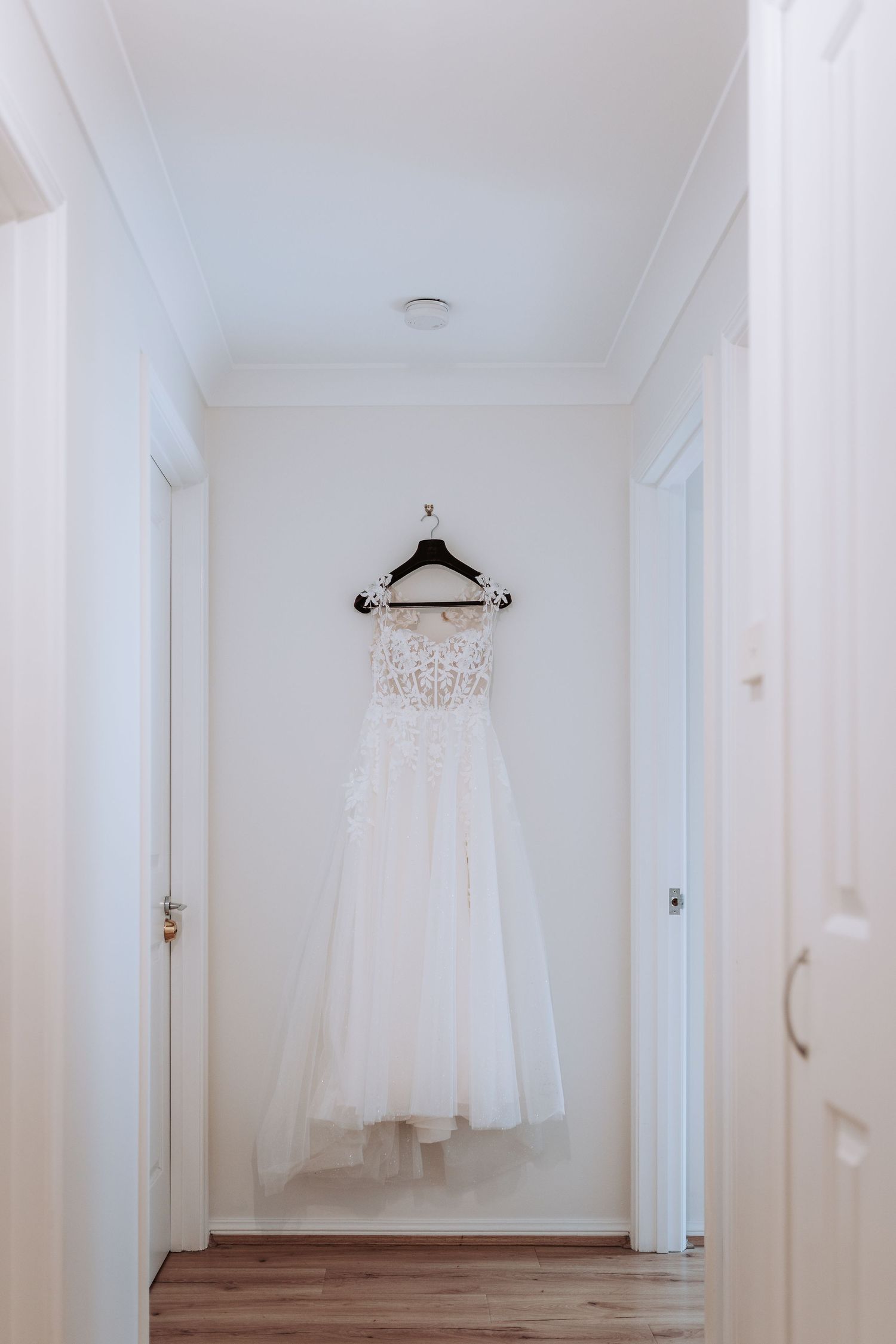 A white wedding dress with lace detailing hangs on a wooden hanger in a minimalist white hallway.