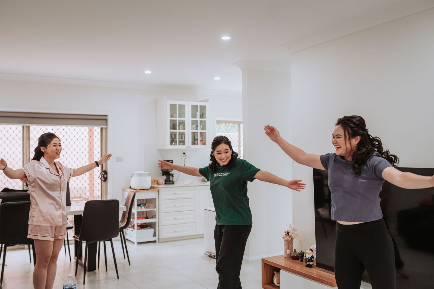 Three people dance joyfully with outstretched arms in a bright, modern living room with white walls and furniture.