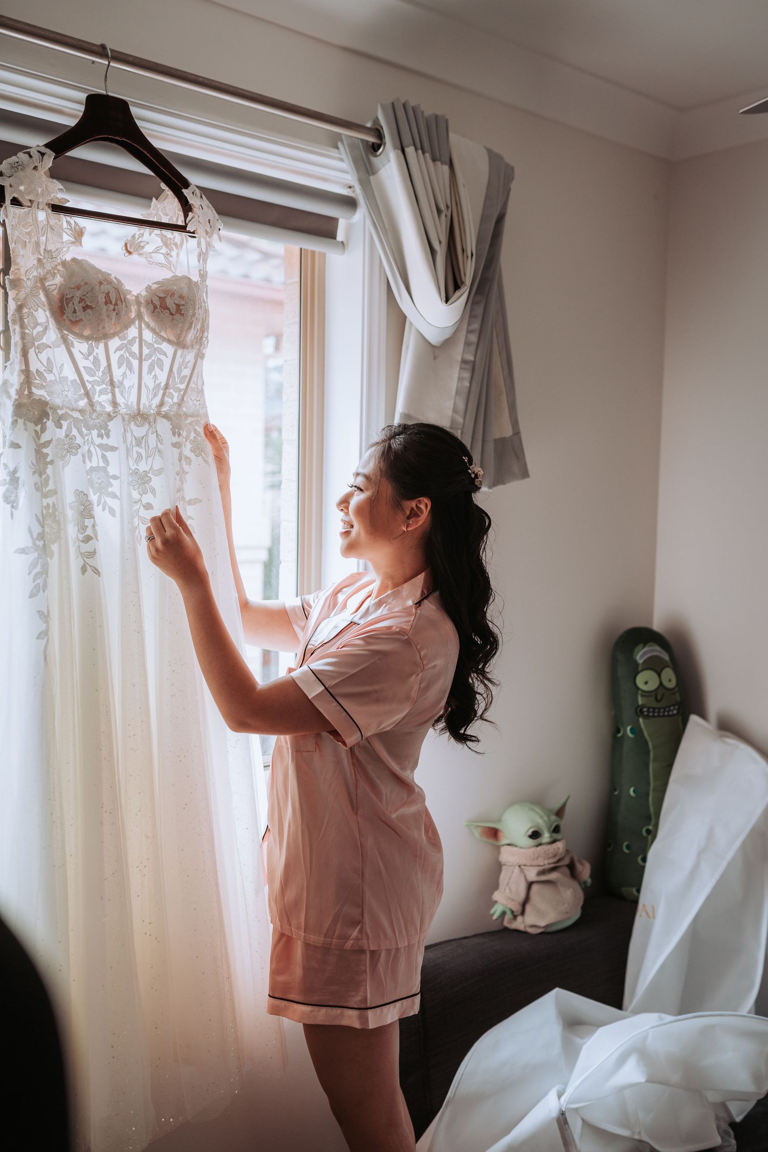 Someone wearing pink satin pajamas admires a white lace wedding dress hanging by a window.