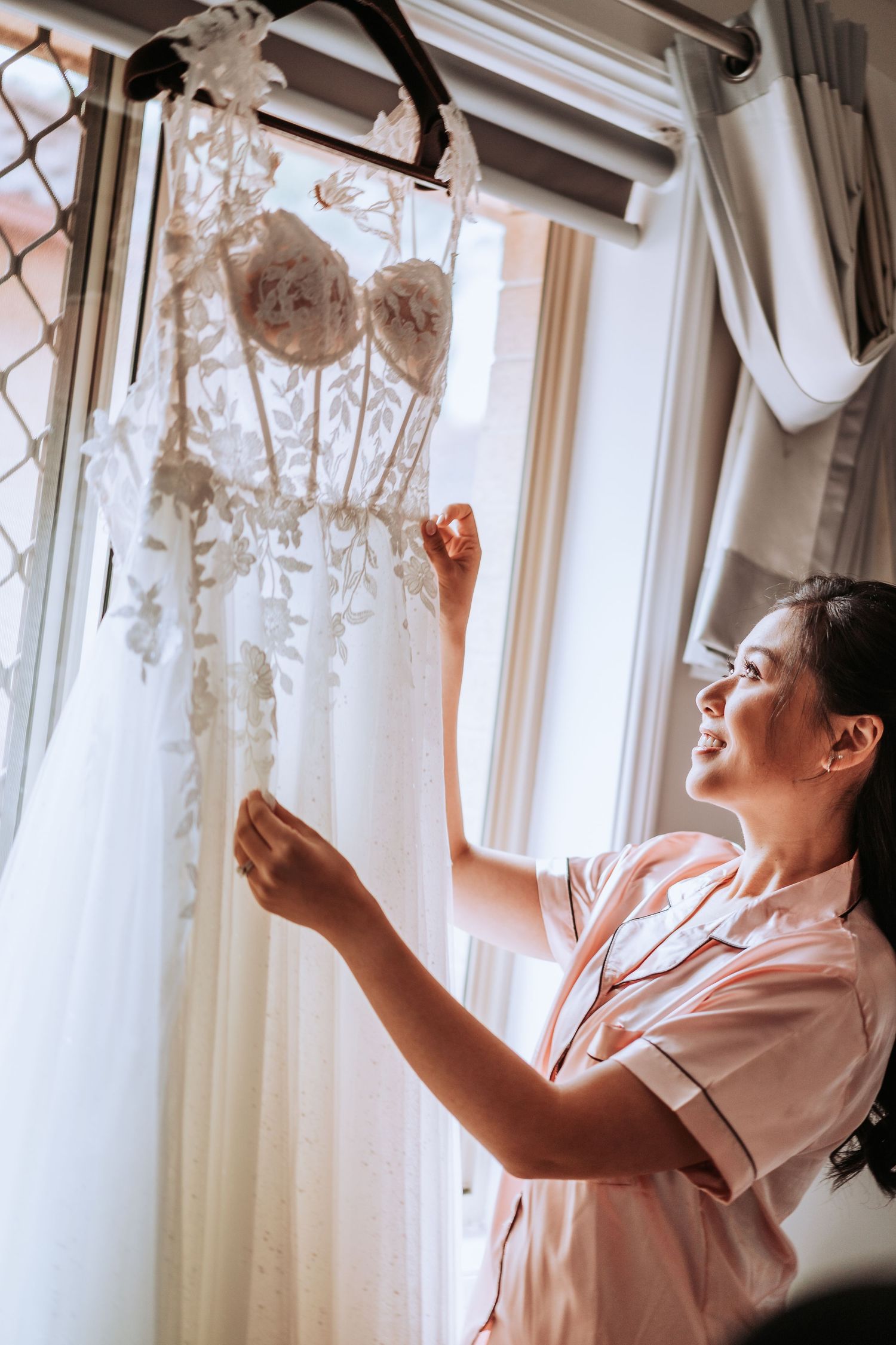 A delicate white lace wedding dress hangs by a window as someone in a pink robe reaches up to touch it.