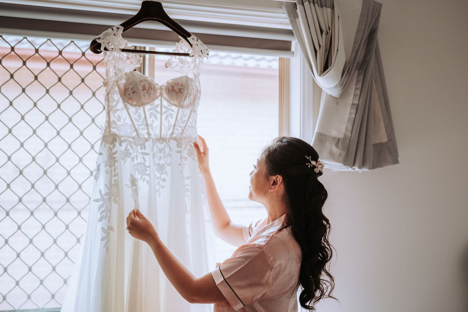 A white lace wedding dress hangs by a window as someone in a robe reaches up to touch it on a hanger.