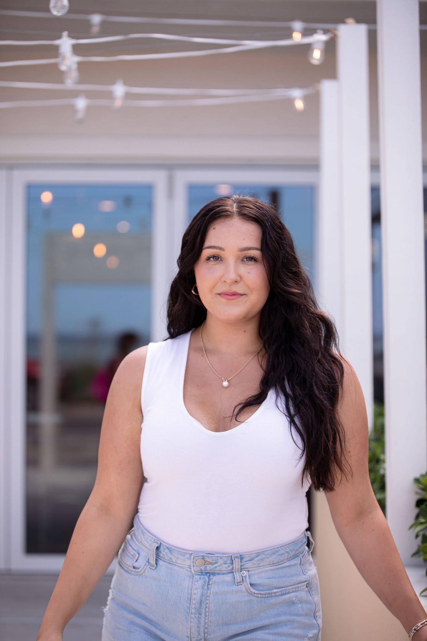 Person in white tank top and jeans standing in front of a building with string lights.