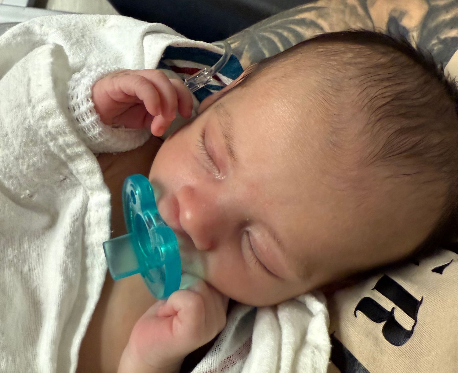 A sleeping newborn baby with a teal-colored pacifier wearing a white outfit.