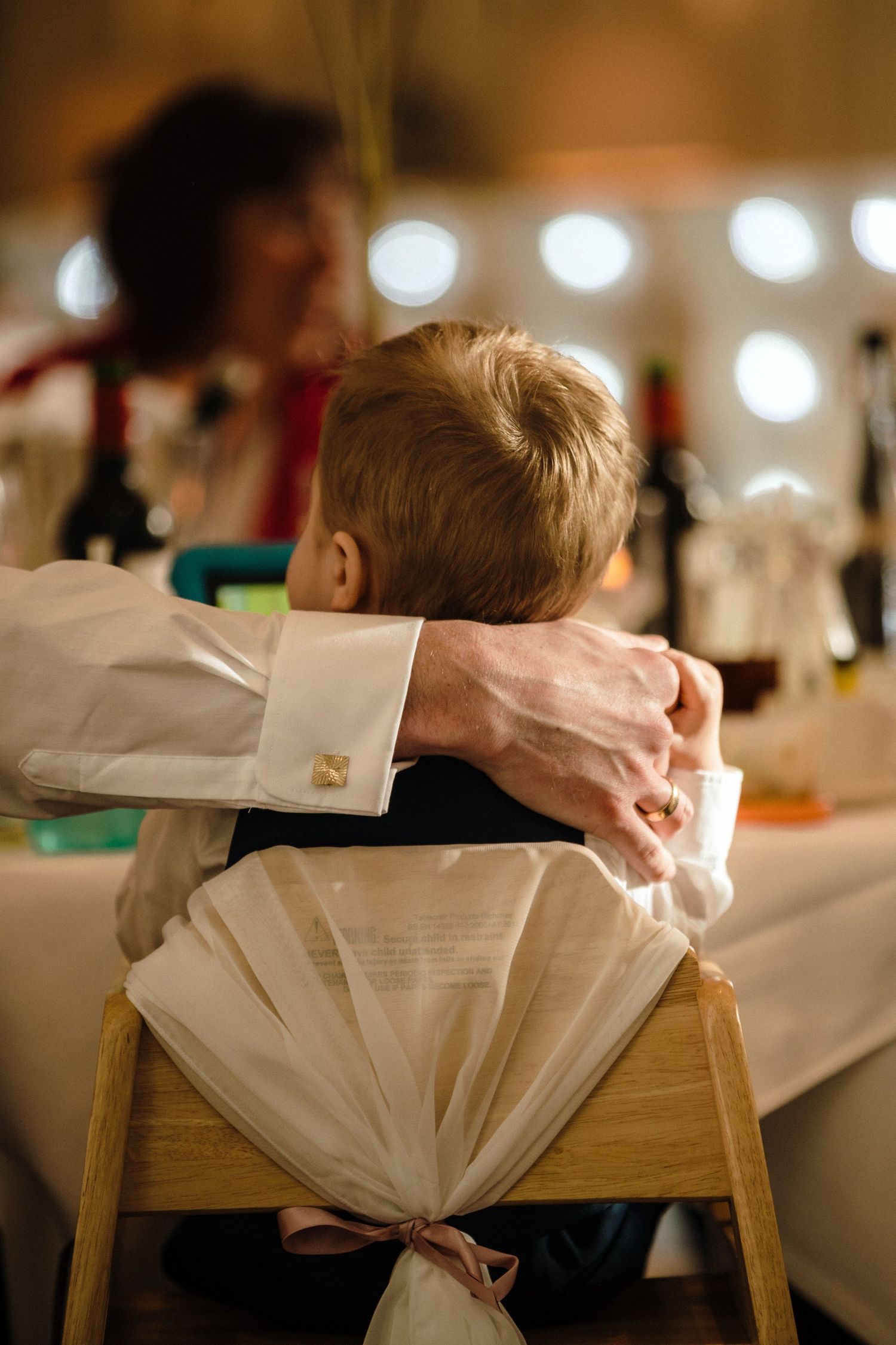 A young child sits in a decorated chair at a formal event, with white fabric and soft lighting in the background.