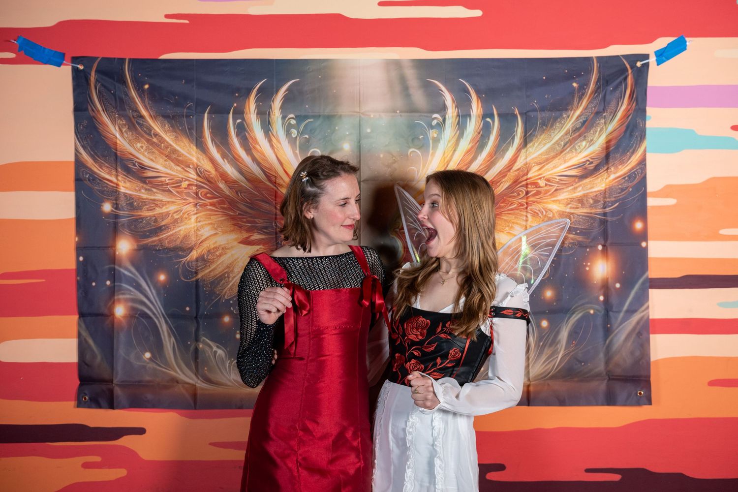 People celebrate in front of dramatic sunset mural with painted angel wings in background.