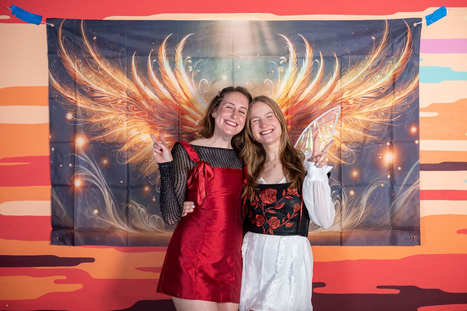 Two people stand against a dramatic wall mural featuring fiery sunset colors and golden wing designs.