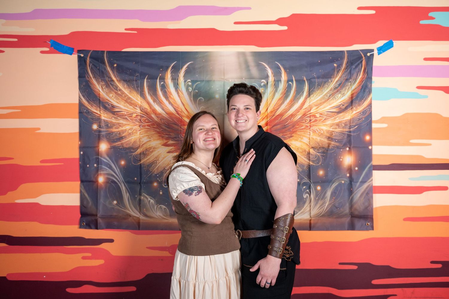 Couples pose dramatically against backdrop featuring glowing wings and sunset colors.